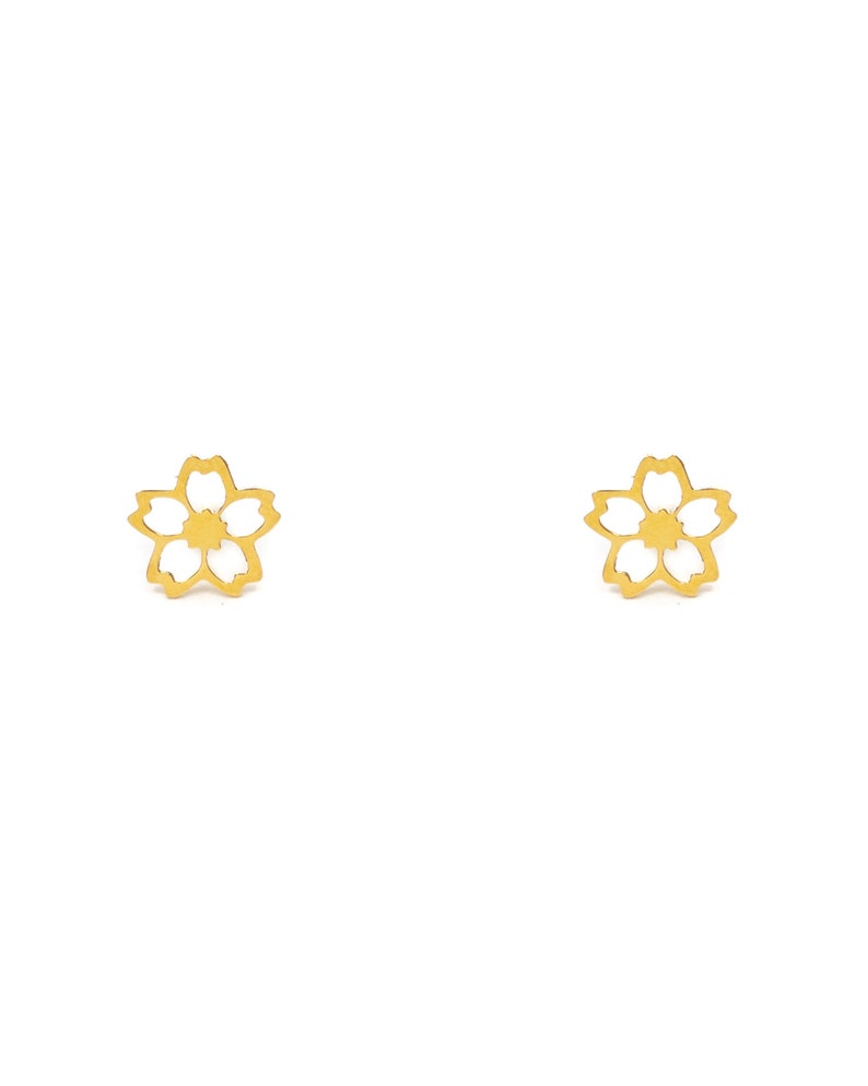 May include: A pair of gold-coloured flower-shaped stud earrings. Each earring features a five-petal design with a cutout centre. The earrings are displayed against a white background.