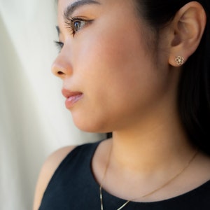 May include: A gold necklace featuring a flower pendant and matching flower stud earrings. The jewellery is worn by a person in a black top. The flower design has six petals and a small central detail. The jewellery is delicate and elegant.