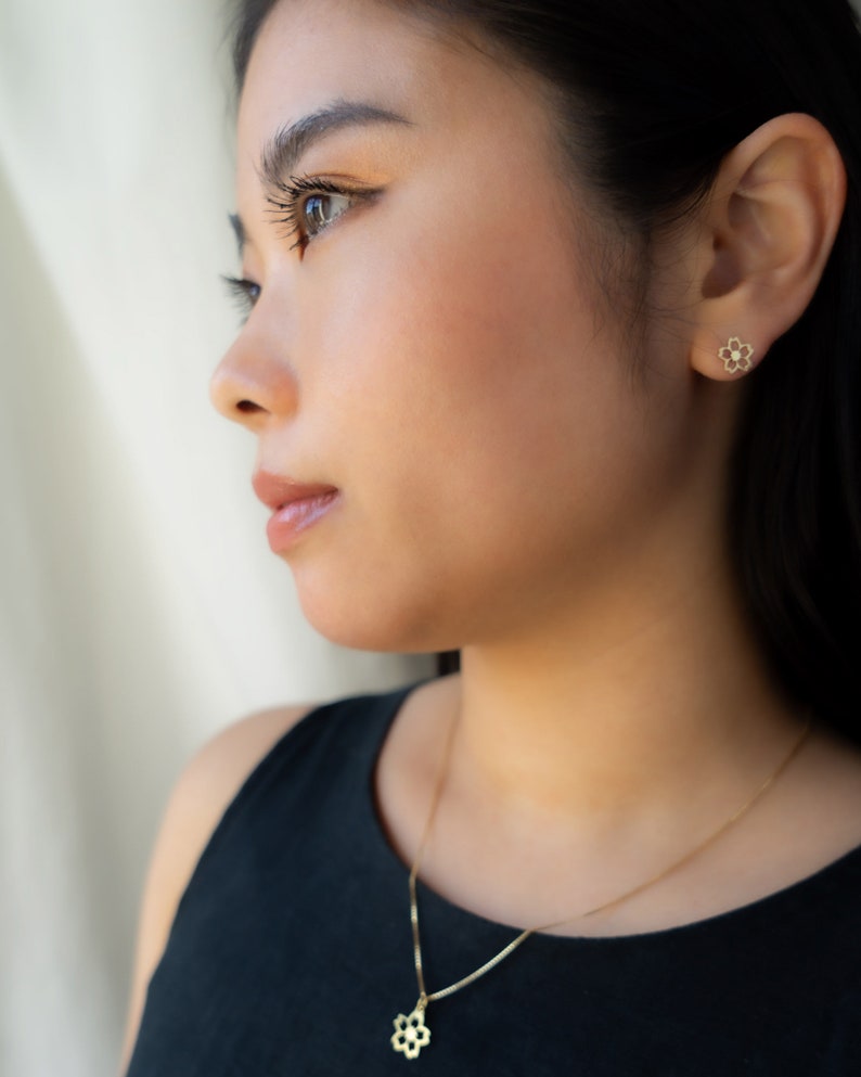 May include: A gold necklace featuring a flower pendant and matching flower stud earrings. The jewellery is worn by a person in a black top. The flower design has six petals and a small central detail. The jewellery is delicate and elegant.