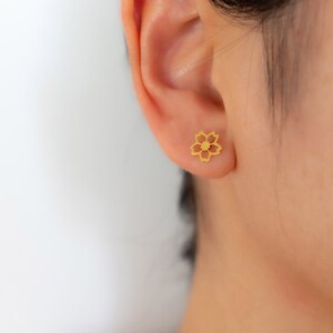 May include: A gold-coloured flower-shaped stud earring is shown on an ear. The earring has a delicate, openwork design with five petals and a central detail. The skin tone is light, and the background is plain white.