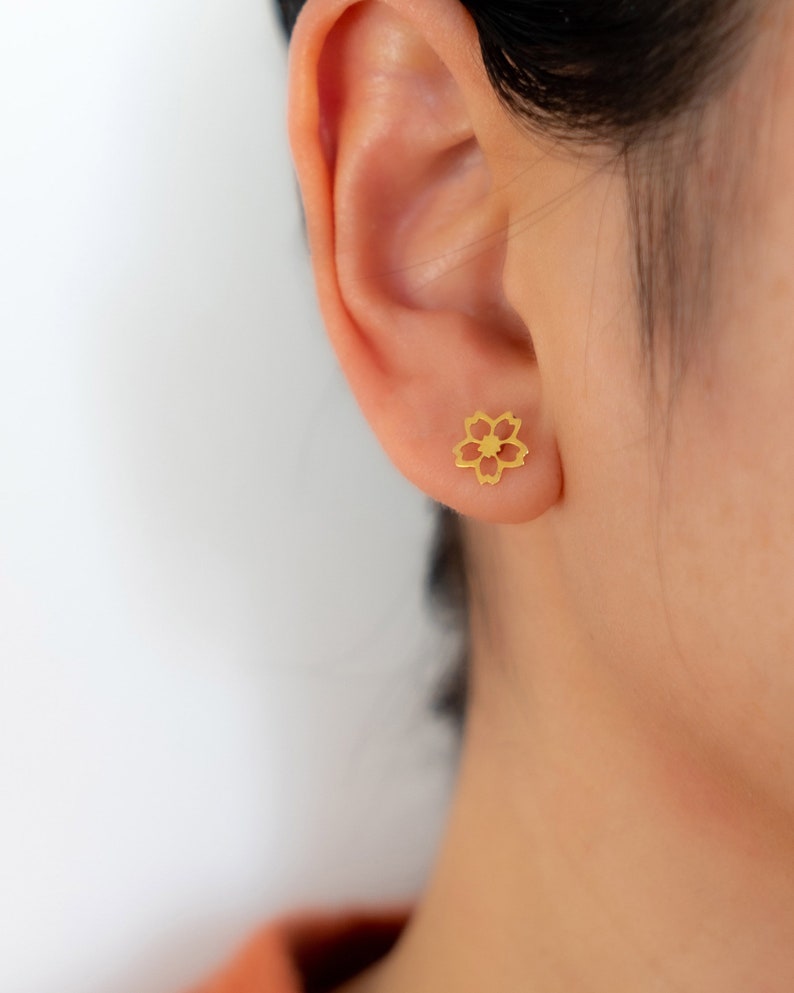 May include: A gold-coloured flower-shaped stud earring is shown on an ear. The earring has a delicate, openwork design with five petals and a central detail. The skin tone is light, and the background is plain white.