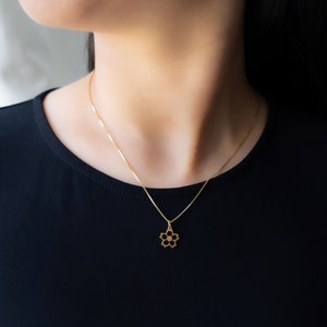 May include: A delicate gold necklace with a five-petal flower pendant. The pendant hangs from a fine chain, offering a minimalist and elegant appearance. The necklace is worn against a black top.