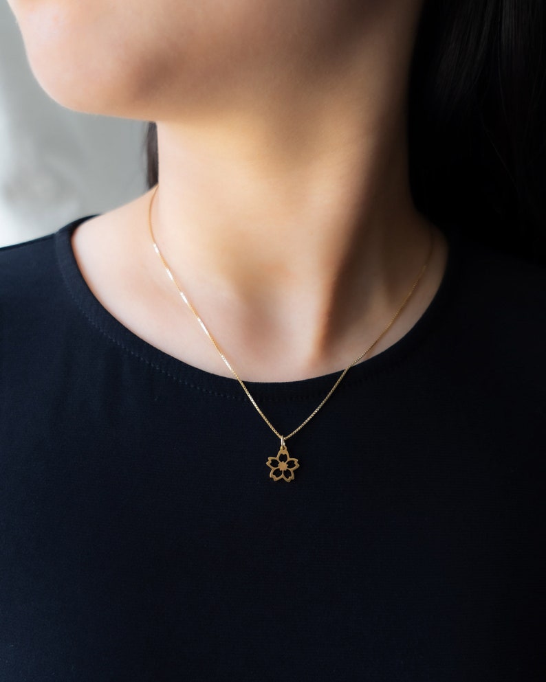 May include: A delicate gold necklace with a five-petal flower pendant. The pendant hangs from a fine chain, offering a minimalist and elegant appearance. The necklace is worn against a black top.