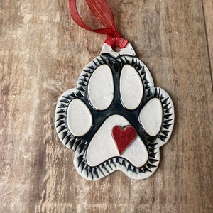 May include: White ceramic paw print ornament with a black outline and a red heart in the center. The ornament has a red ribbon attached for hanging.