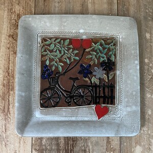May include: A white ceramic square plate with a raised border. The center of the plate features a brown tile with a black bicycle, red balloons, and flowers. The tile is surrounded by a white border with a decorative pattern. The plate has a red heart and the word "Teal" written in cursive.