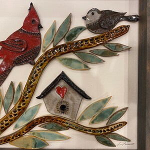 May include: A ceramic wall art piece featuring a red cardinal, a small brown bird, and a birdhouse with a red heart on a brown branch with green leaves.