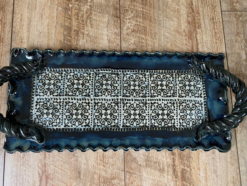 May include: Rectangular ceramic serving tray with a dark blue glaze and a decorative white and black tile pattern. The tray has a wavy edge and twisted rope handles, perfect for serving appetizers or desserts.