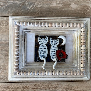 May include: A white framed ceramic tile with two white cats with black spots, a red poppy, and a crescent moon. The cats are facing away from the viewer and their tails are curled up.