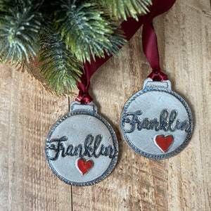 May include: Two white ceramic Christmas ornaments with blue trim and the name "Franklin" written in black script. Each ornament has a red heart painted on it.