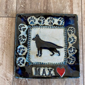 May include: A square ceramic tile with a blue and white glaze. The tile features a silhouette of a German Shepherd dog, paw prints around the border, and the name "MAX" in black lettering. A red heart is in the bottom right corner.