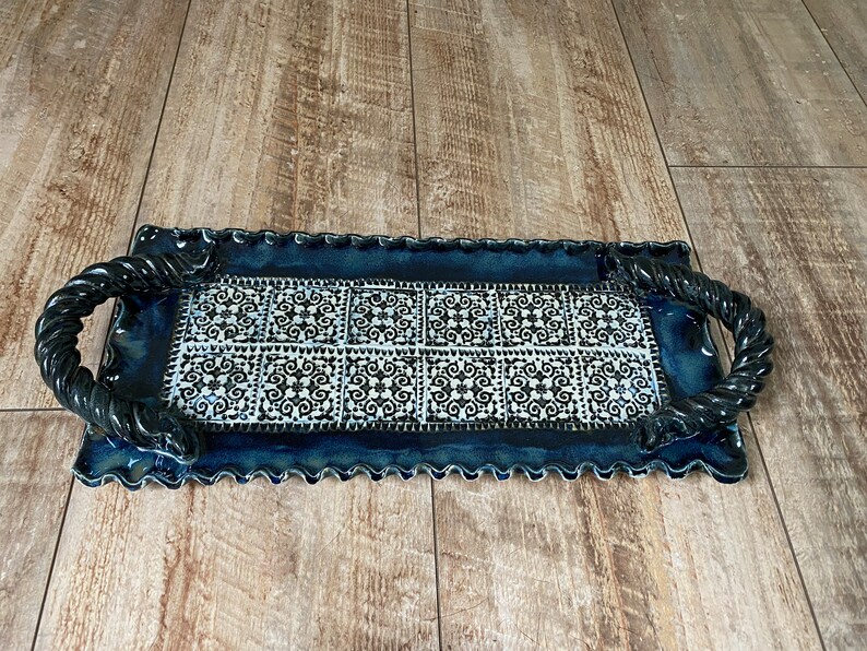 May include: Rectangular ceramic serving tray with a dark blue glaze and a decorative black and white tile pattern. The tray has twisted rope-style handles and a scalloped edge. A decorative piece for serving.