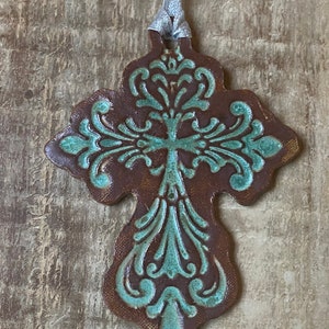 May include: A brown ceramic cross ornament with a teal blue design. The cross has a decorative, ornate design and is hanging from a silver ribbon.