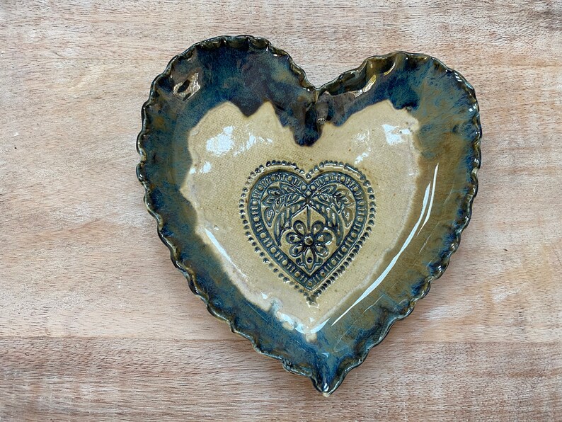 Puede incluir: Un plato de cer&aacute;mica en forma de coraz&oacute;n con un borde festoneado. El plato est&aacute; esmaltado en color azul y marr&oacute;n con un dise&ntilde;o de coraz&oacute;n marr&oacute;n m&aacute;s claro en el centro.