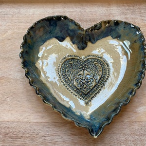 Puede incluir: Un plato de cer&aacute;mica en forma de coraz&oacute;n con un borde festoneado. El plato est&aacute; esmaltado en azul y marr&oacute;n con un interior blanco. Un dise&ntilde;o m&aacute;s peque&ntilde;o en forma de coraz&oacute;n est&aacute; en relieve en el centro del plato.