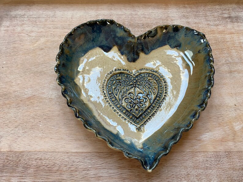 Puede incluir: Un plato de cer&aacute;mica en forma de coraz&oacute;n con un borde festoneado. El plato est&aacute; esmaltado en azul y marr&oacute;n con un interior blanco. Un dise&ntilde;o m&aacute;s peque&ntilde;o en forma de coraz&oacute;n est&aacute; en relieve en el centro del plato.