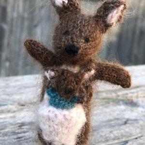 Puede incluir: Una figura de canguro tejida en color marrón difuso con orejas largas que presentan detalles interiores en rosa claro. El canguro tiene una bolsa que contiene una pequeña cría tejida en rosa claro y una pieza decorativa azul cerceta.