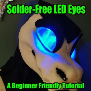 Könnte beinhalten: Nahaufnahme einer weißen Fellmaske mit blauen und schwarzen Akzenten. Die Maske hat zwei blaue LED-Augen, die leuchten. Der Text "Solder-Free LED Eyes" ist in grünen Buchstaben oben im Bild geschrieben. Der Text "A Beginner Friendly Tutorial" ist in grünen Buchstaben unten im Bild geschrieben.