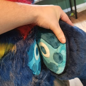 Può includere: Una coda pelosa blu e verde con un'apertura a cerniera. La coda è tenuta da una mano. Il testo "How to make a Zipper Tail" e "PDF Tutorial" è visibile nell'immagine.
