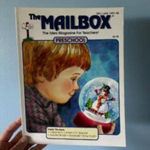 May include: The Mailbox magazine cover featuring a young child looking at a snow globe with a snowman inside. The magazine title is in blue and the word "Preschool" is in a yellow box. The background is light blue.