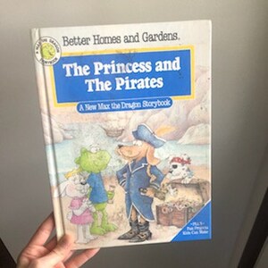May include: A children's book titled "The Princess and The Pirates" with a blue cover and illustrations of a dragon, a princess, and a pirate. The book is part of the "Max the Dragon Storybook" series.