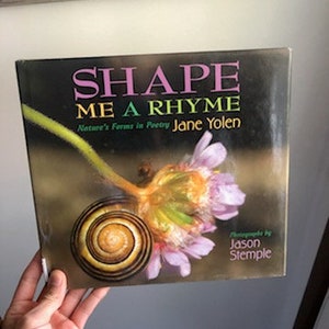 May include: A book titled "Shape Me a Rhyme: Nature's Forms in Poetry" by Jane Yolen. The cover features a close-up of a pink flower with a spiral-shaped snail shell.