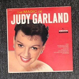 May include: A red vinyl record album cover featuring a portrait of Judy Garland. The album title is "The Magic of Judy Garland" with the Decca Records logo in the top right corner.