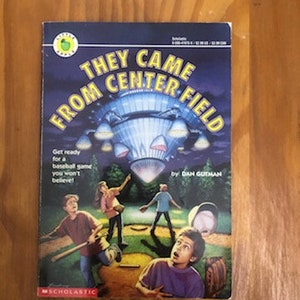 May include: A Scholastic book cover with the title "They Came From Center Field" and a picture of a flying saucer over a baseball field. The book is by Dan Gutman.