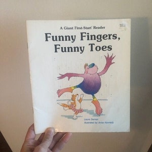 May include: A children's book titled "Funny Fingers, Funny Toes" with an illustration of a purple monster with a yellow and orange striped shirt and a small orange and white dog.