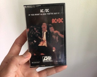May include: A black cassette tape with a white label. The label has the text "AC/DC" and "IF YOU WANT BLOOD YOU'VE GOT IT" in black text. The band's logo is in red and white. The cassette tape is labeled "CS 19212".