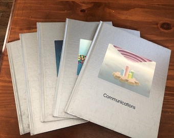 May include: A stack of silver-colored books with the word "Communications" printed on the cover. The cover features a colorful graphic of a city on a cloud. The books are on a wooden surface.