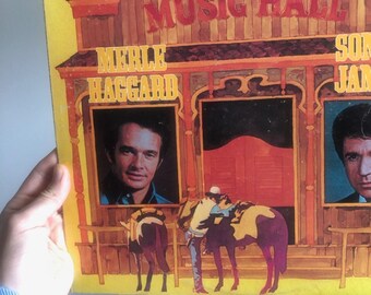 May include: A yellow vinyl record album cover with a vintage illustration of a wooden music hall building. The album features Merle Haggard and Sonny James. The text on the cover reads "Plaza House presents Music Hall".