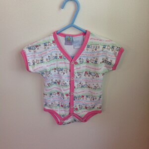 May include: A white baby onesie with a pink trim and a button closure. The onesie has a colorful pattern of animals and stripes. The onesie is hanging on a blue hanger.