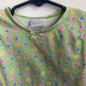 May include: A green short-sleeved shirt with a small white flower print. The shirt has a green trim around the neckline and a white tag that says "Carter's Since 1965".