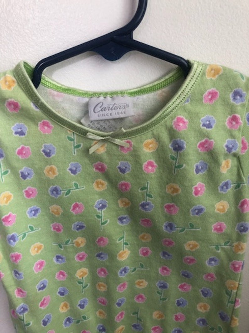 May include: A green short-sleeved shirt with a small white flower print. The shirt has a green trim around the neckline and a white tag that says "Carter's Since 1965".