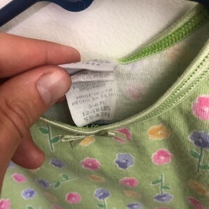 May include: Green sleeveless dress with a floral print featuring pink, yellow, and purple flowers. The dress has a tag that reads "Made in USA, Decor, 12-18 lbs, 5.5-8.2kg."