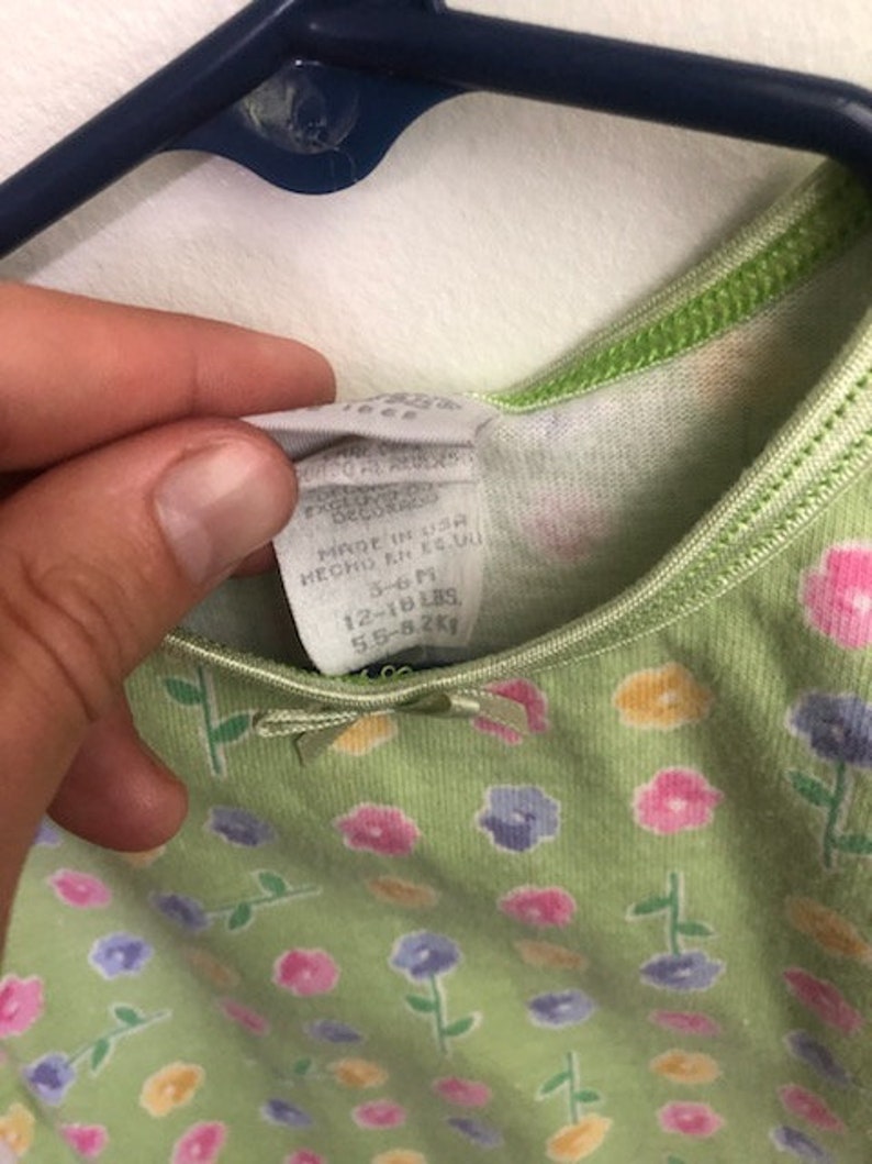 May include: Green sleeveless dress with a floral print featuring pink, yellow, and purple flowers. The dress has a tag that reads "Made in USA, Decor, 12-18 lbs, 5.5-8.2kg."