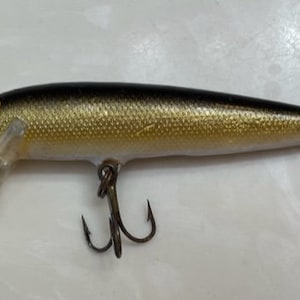 May include: A gold and black fishing lure with a single hook and a clear plastic fin.