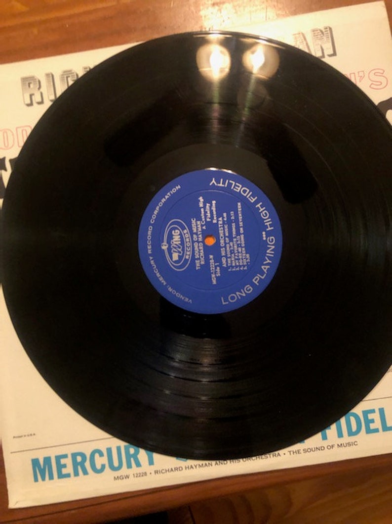 K&ouml;nnte beinhalten: Eine schwarze Schallplatte mit einem blauen Etikett, auf dem "Long Playing High Fidelity" und "The Sound of Music" steht, mit dem K&uuml;nstler Richard Hayman und seinem Orchester. Die Schallplatte befindet sich auf einem wei&szlig;en Hintergrund mit dem Text "Mercury Records Corporation" und "MGW 12228".