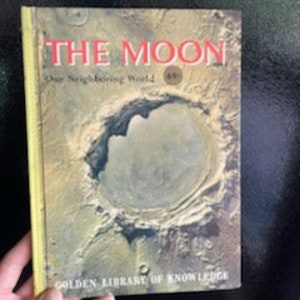 May include: A vintage hardcover book titled "The Moon Our Neighboring World" with a golden cover and a large image of a crater on the moon. The book is part of the Golden Library of Knowledge series.