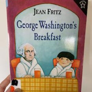 May include: A pink book with a yellow and white illustration of George Washington and a boy sitting at a table with orange juice. The book title is "George Washington's Breakfast" by Jean Fritz.