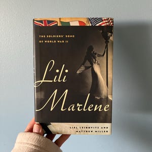 May include: A hardcover book titled Lili Marlene: The Soldiers' Song of World War II by Liel Leibovitz and Matthew Miller. The cover features a silhouette of a person in a gown standing by a lamp post, with national flags displayed at the top.