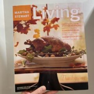 May include: A hand holds a copy of Martha Stewart Living magazine. The cover features a roasted turkey on a platter with vegetables. The magazine title "Living" is in large white letters, with "Thanksgiving" at the bottom.