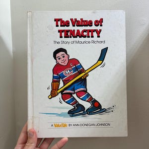 May include: A hardcover book titled The Value of Tenacity: The Story of Maurice Richard. The white cover features a cartoon illustration of a hockey player in a red, white, and blue uniform holding a yellow hockey stick on the ice. The text A ValueTale by Ann Donegan Johnson is printed at the bottom.