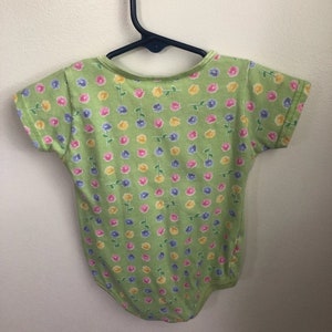 May include: A green baby onesie with a floral print featuring pink, yellow, and blue flowers. The onesie has short sleeves and a snap closure.