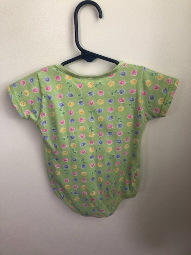 May include: A green baby onesie with a floral print featuring pink, yellow, and blue flowers. The onesie has short sleeves and a snap closure.