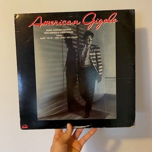 May include: A black vinyl record album cover with the title "American Gigolo" in red lettering. The cover features a man in a striped suit standing in a room with blinds. The text on the cover reads "Original Soundtrack Recording, Music Composed by Giorgio Moroder, Featuring, Blondie, "Call Me",  Cheryl Lynn, "Love & Passion"