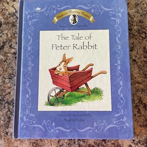May include: A blue book with a gold border and a picture of a rabbit in a wheelbarrow. The title is "The Tale of Peter Rabbit" and the book is by Beatrix Potter.