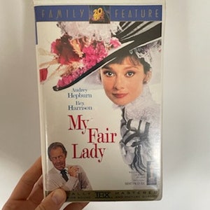 May include: A VHS tape of the movie My Fair Lady starring Audrey Hepburn and Rex Harrison. The cover features a photo of Audrey Hepburn in a large hat with flowers and a white dress. The title of the movie is in red and white text.