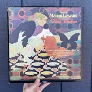 May include: A vintage vinyl record album cover featuring an illustration of a woman and a man in period clothing. The album title "Manon Lescaut" is visible, along with the names of the performers. The cover has a colorful, patterned background.