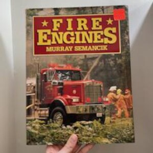May include: A book titled "FIRE ENGINES" by Murray Semancik. The cover features a red fire truck spraying water, with firefighters in orange suits. The background shows a forest scene.